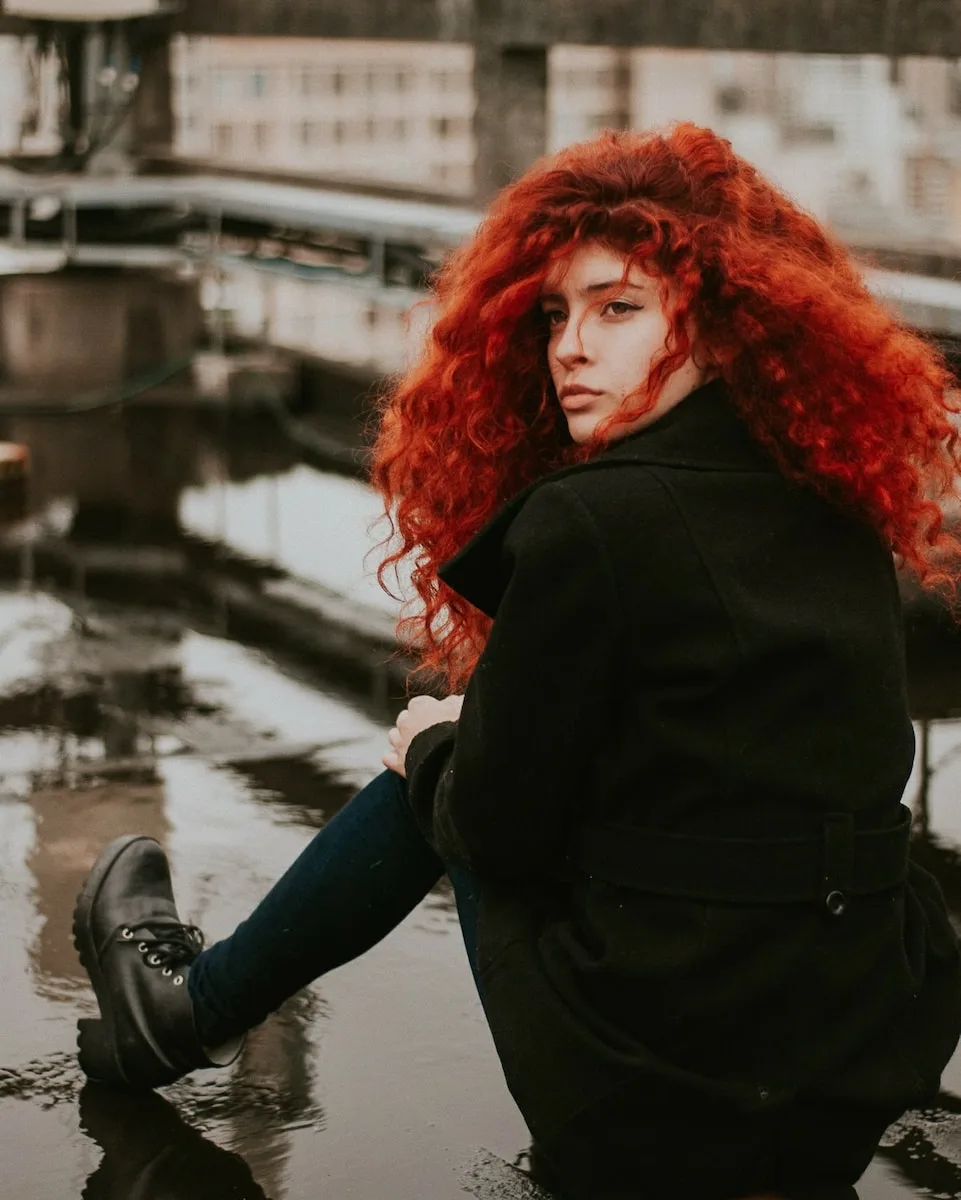 woman with vibrant red hair sitting in an urban waterfront setting, looking over her shoulder
